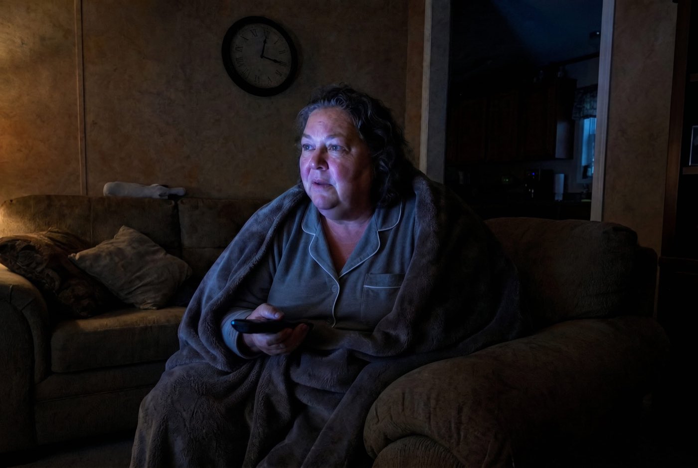 A woman ~55 sitting alone on her living-room sofa in the dark at night, wrapped in a soft blanket, lit only by the blue glow of a TV. Her face is tired but suddenly alert and leaning forward, remote in hand, fully caught by what she's watching. A clock faintly reads about 3:00.